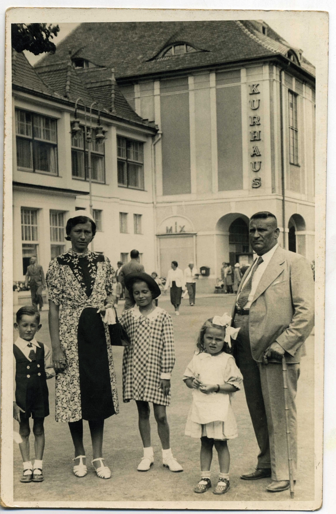 Zdjęcie rodziny. Po lewej stronie kobieta trzyma dzieci, dziewczynkę i chłopca, za ręce. Po prawej stronie mężczyzna stoi o lasce obok niego stoi dziewczynka.