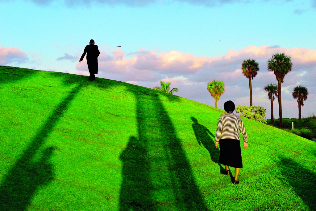Dwoje ludzi, kobieta i mężczyzna idą wzgórzem. Kobieta jest niżej, a mężczyzna wyżej. Na trawie odbijają się cienie.