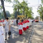 Procesja Bożego Ciała idzie ulicami miasta, obok ludzie przejeżdża tramwaj.