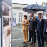 Czworo ludzi stoi, trzech mężczyzn i kobieta oglądają wystawę plenerową. Dwóch mężczyzn trzyma parasole.