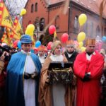 Na pierwszym planie trzech mężczyzn ubranych w stroje królów. Na drugim planie ludzie z flagami i balonami.