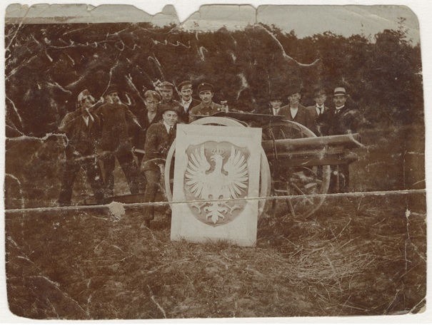 Grupa mężczyzn stoi za działem paryskim, przed nim ustawione godło z orłem w koronie.
