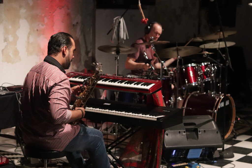dwóch mężczyzn siedzących w otoczeniu instrumentów muzycznych, jeden gra na perkusji, drugi trzyma w ręce saksofon