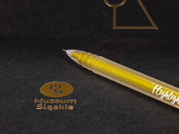 Mocne zbliżenie na czarną okładkę notesu. Widoczna faktura oraz tłoczenie logo Muzeum i logo wystawy w kolorze złotym. Na okładce leży żelopis, również w kolorze złota.