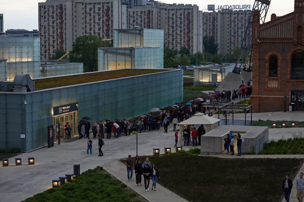 Budynki z przeszklonymi ścianami i zielonym dachem. Wokół budynku widać wiele osób. W tle bloki mieszkalne.