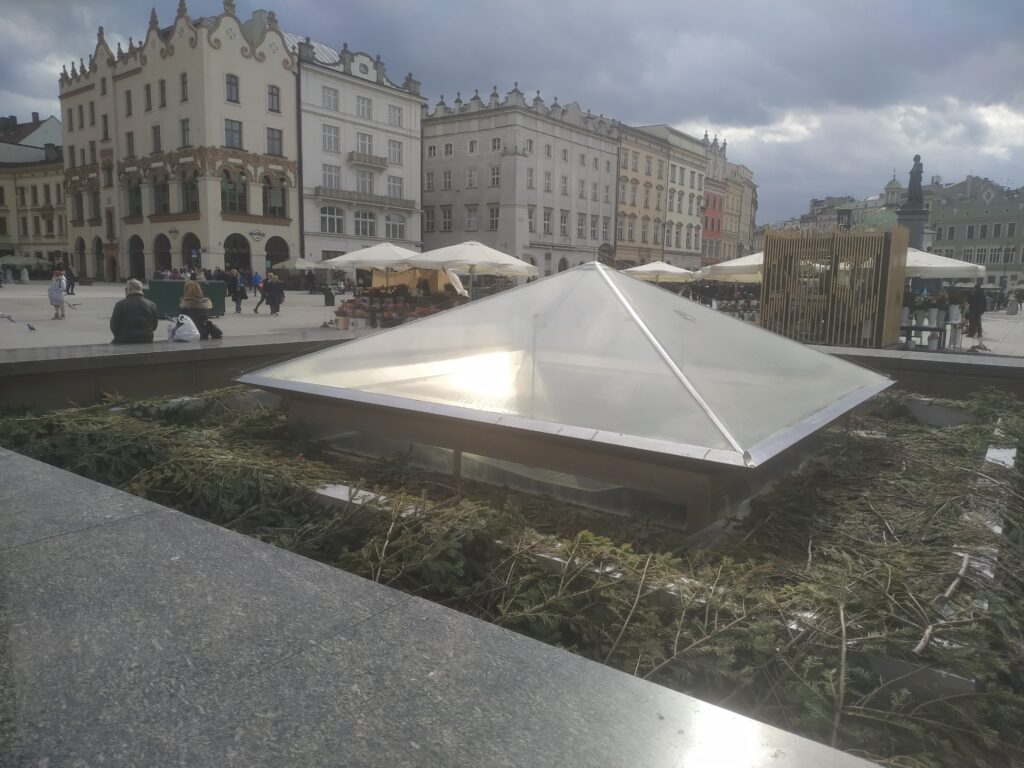 Szklana piramida na tle krakowskiego placu, otoczona zielonymi gałęziami. W tle są kamienice i przechodnie.