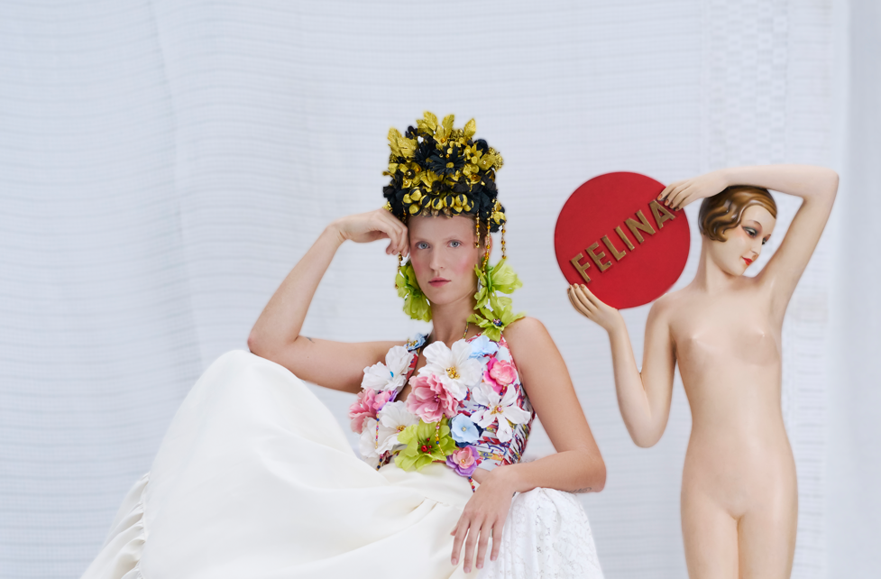 Zdjęcie z sesji zdjęciowej promującej wystawę "Etnoalternatywa". Na białym tle kobieta w białej sukni z kwiatami, obok figura nagiej kobiety trzymającej napis "FELINA"