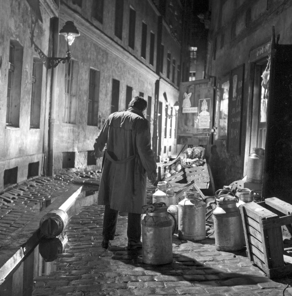 czarno-białe zdjęcie, mężczyzna w jasnym płaszczu stoi sam na ulicy, dookoła niego leżą porozrzucane palety, świecą uliczne latarnie