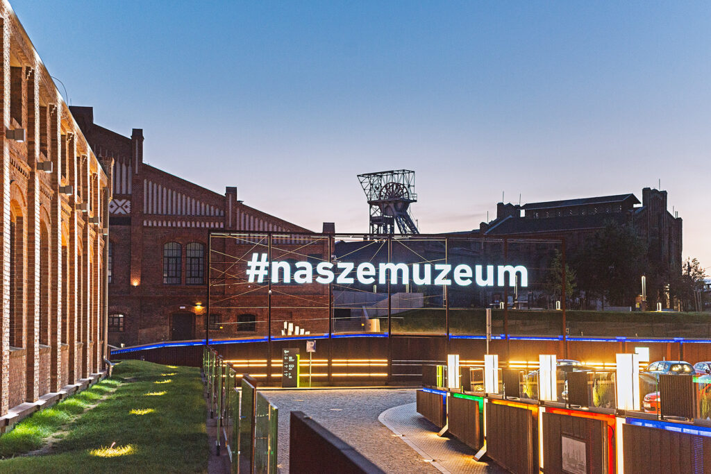 Teren Muzeum Śląskiego w Katowicach o zmierzchu. W centralnym punkcie duży, świecący biały neon z napisem „#naszemuzeum”, a w tle zabytkowe ceglane budynki i wieża szybu kopalnianego.