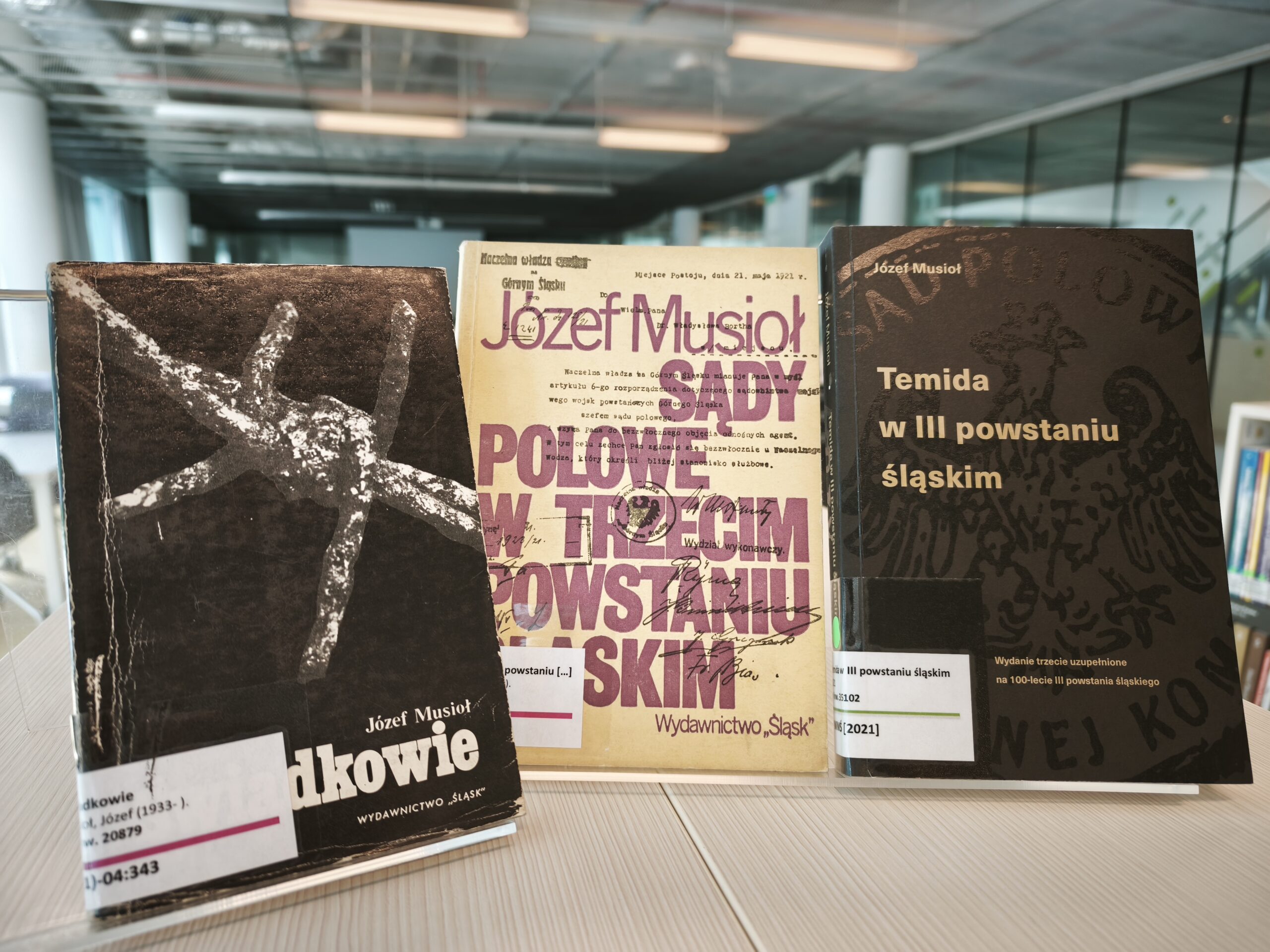 trzy książki stojące na białym blacie, na każdej z nich widać naklejkę Muzealnej Biblioteki